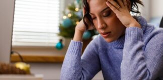 6 Key Strategies for Lowering Your Stress Levels This Holiday Season A person with curly brown hair in a ponytail and light purple sweater holding their head with their hands at their desk in front of a small Christmas tree with blue ornaments and white lights.