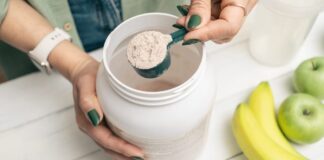 Collagen: Considering Its Health Benefits Beyond the Anti-Aging Claims A person with green nails and a smartwatch scooping a white collagen powder out of a container next to a banana and two green apples.
