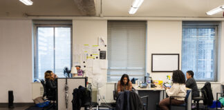 Students Collaborate with Toronto on Mental Health Response Students sit around a table and at a desk in a classroom. Some are on the phone and some are working on their laptops.