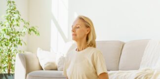 The Science Behind Breathwork and Stress Reduction Peaceful senior woman in lotus position meditation with closed eyes at home while sitting on yoga mat on floor, full length.