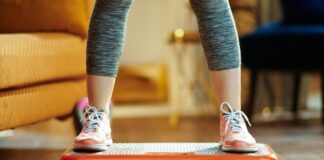 Do Whole Body Vibration Plates Actually Promote Weight Loss? We Asked Fitness Experts A close-up of a person wearing grey leggings and sneakers standing on a vibration plate in a living room with a mustard yellow couch.