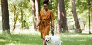 What Personal Trainers Think About Walking Becoming Trendy Again A person with brown hair up in a bun walking a white dog in a park surrounded by trees.