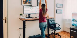 Wellness Devices Recommend by the Fitness Professionals Who Use Them A person in jeans and a dark pink-purple tee using an under-desk treadmill while working on the computer.