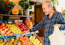 Celebrating March as National Nutrition Month Elderly man in a plaid shirt smiling while selecting fruits from a market stand during National Nutrition Month. Vibrant apples, pears, and tomatoes are displayed in boxes nearby.
