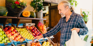 Celebrating March as National Nutrition Month Elderly man in a plaid shirt smiling while selecting fruits from a market stand during National Nutrition Month. Vibrant apples, pears, and tomatoes are displayed in boxes nearby.